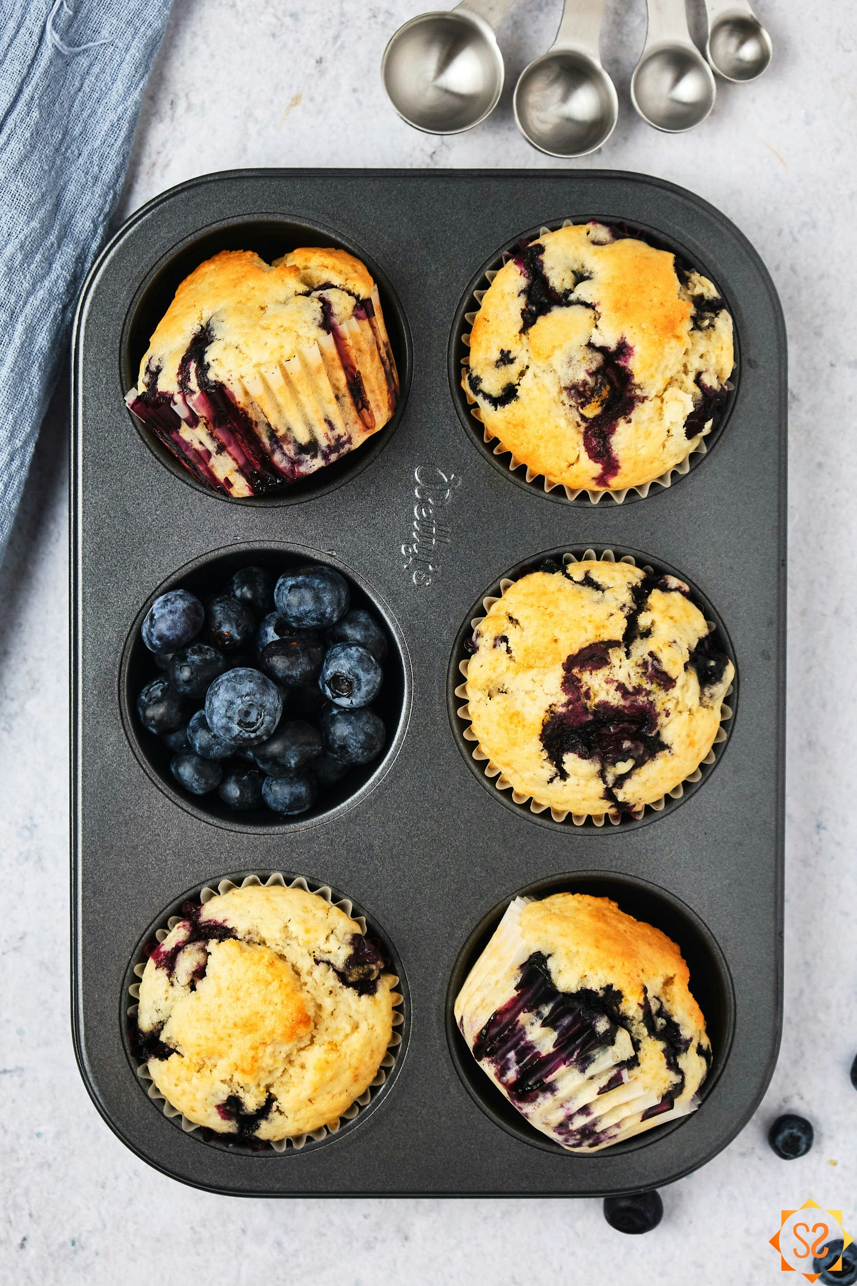 A top-down view of vegan blueberry muffins in a pan, with one hole filled with blueberries, and measuring spoons to the side.