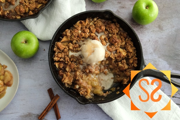 A skillet of apple crisp with vegan vanilla ice cream on top, surrounded by apples and cinnamon sticks, and a plate of apple crisp.