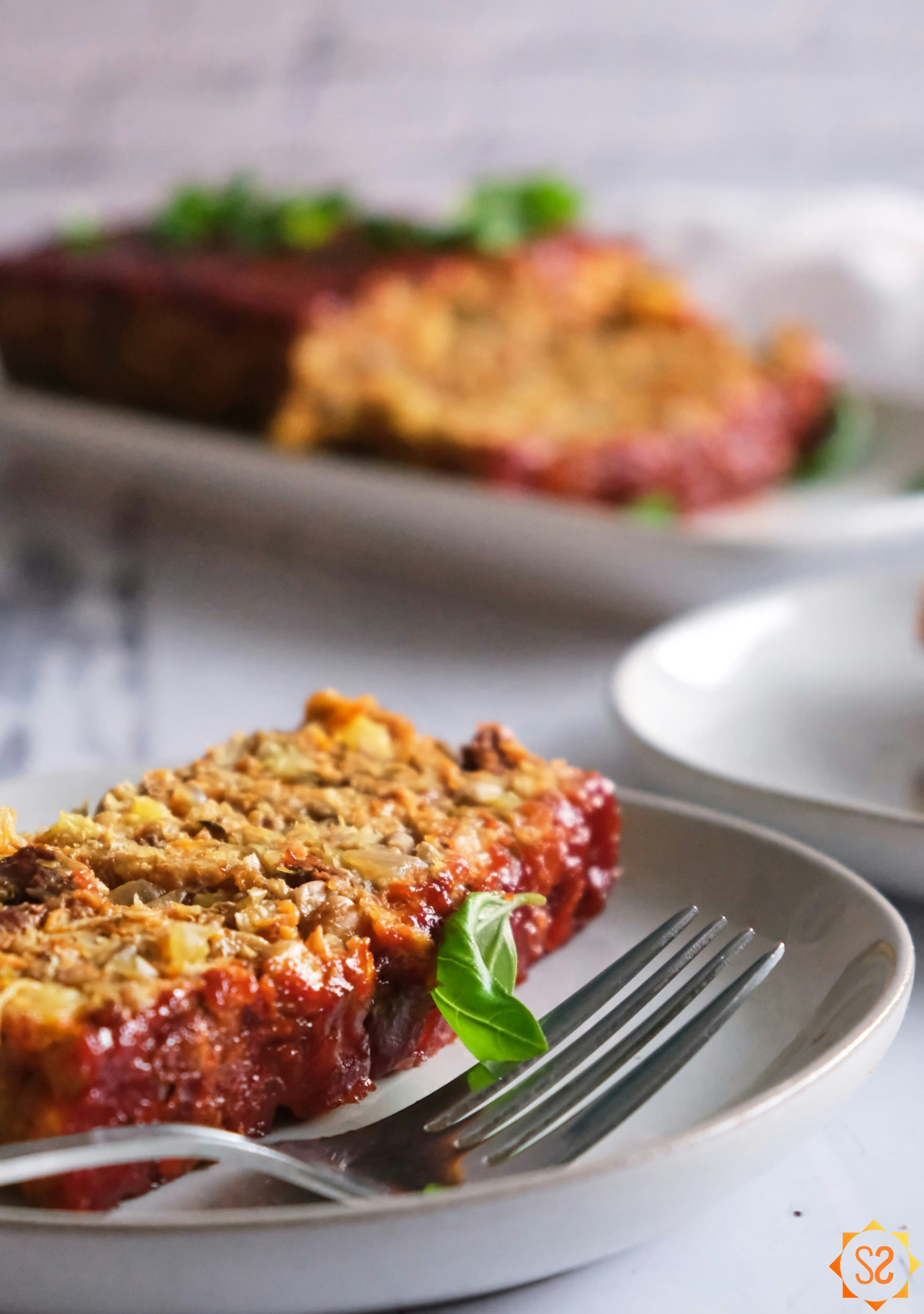 A slice of lentil loaf on a plate with a fork, with the full loaf in the background