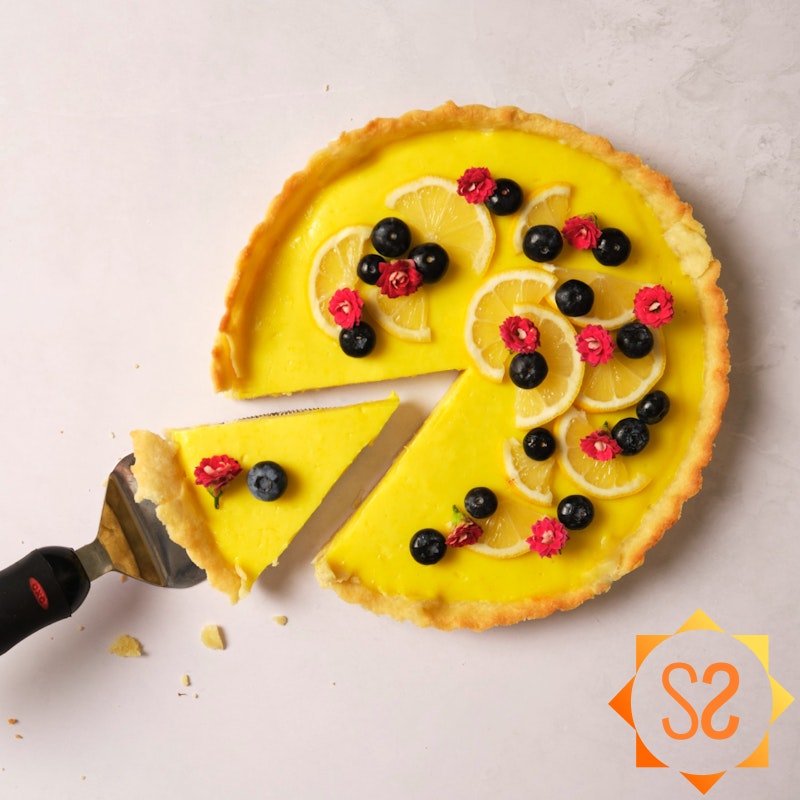 A vegan lemon tart topped with berries, flours, and lemon slices, with one slice being pulled out with a pie server.