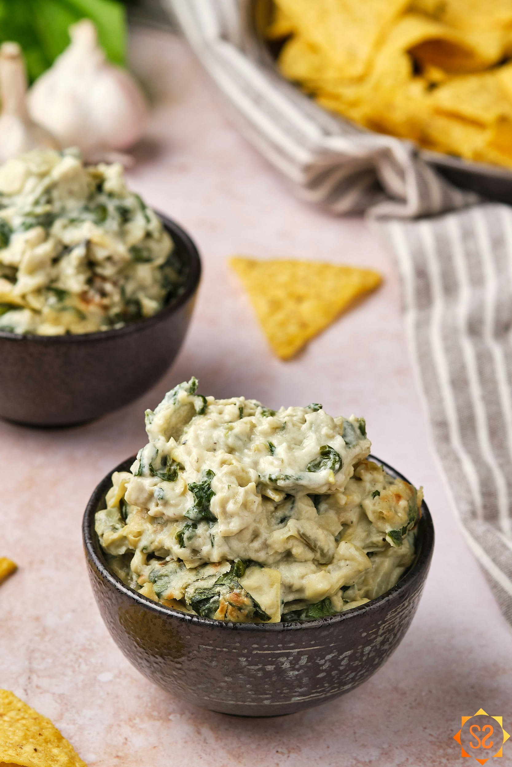 Two bowls of vegan spinach and artichoke dip, with chips in the background.