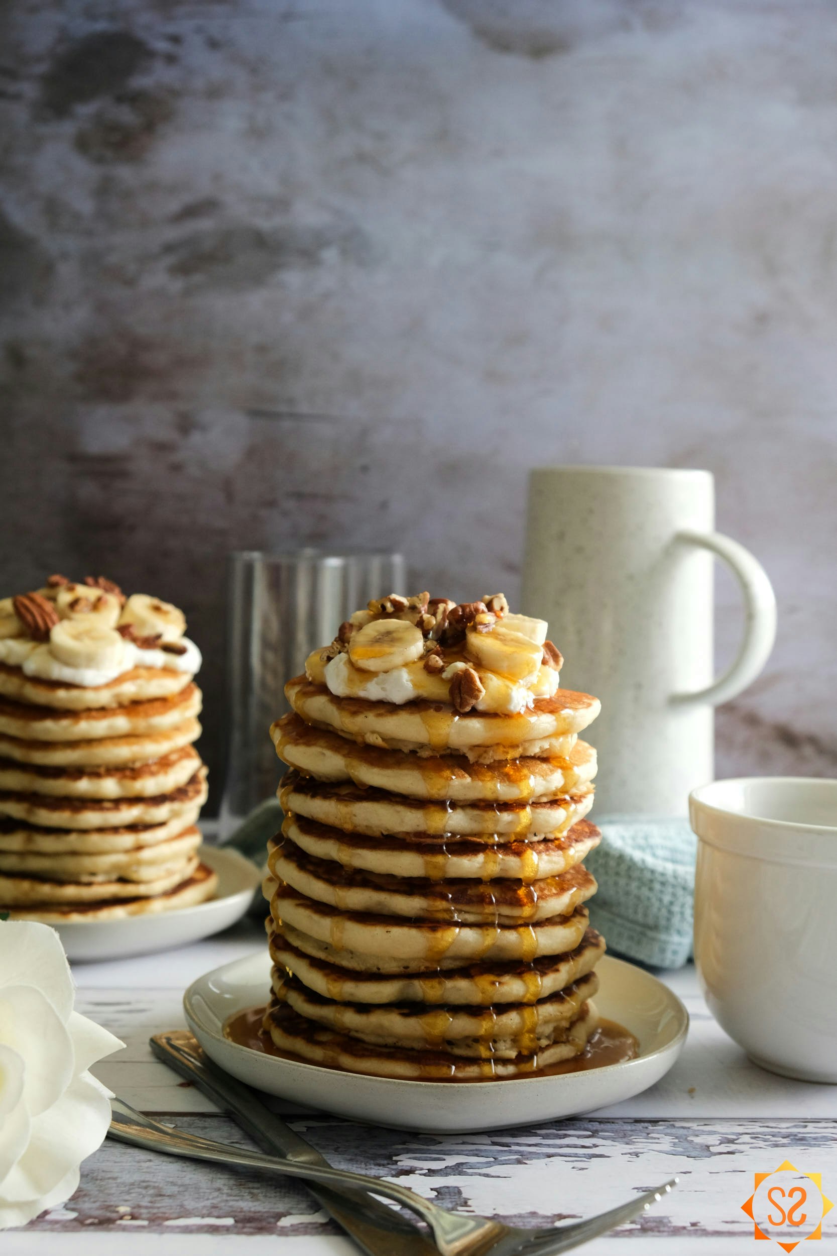 A stack of vegan buttermilk pancakes with maple syrup, whipped cream, bananas, and pecans on top.