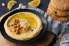 A bowl of hummus with a stack of pitas to the side, lemon wedges and garlic cloves behind it.
