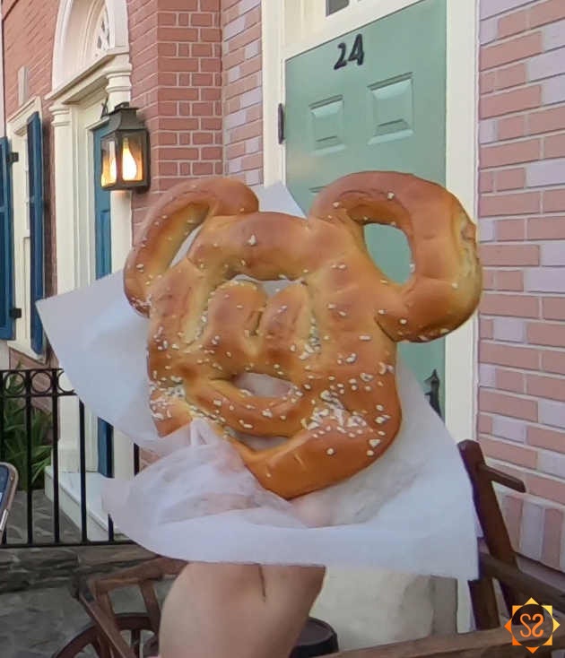 Mickey Pretzel in a hand in front of a Disney townhouse facade.