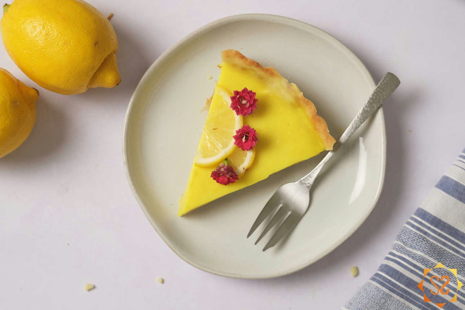A top-down view of a slice of vegan lemon tart on a plate with two lemons to the side.