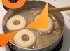 Donuts frying in a pan, with tongs removing a piece of parchment paper from one of the donuts.