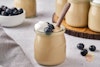 A single jar of vegan vanilla pudding with a spoon, with more jars and blueberries in the background. 