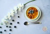 a creme brulee dish topped with blueberries; a spoon is to the bottom of it, more blueberries and an orchid stem are to the left of the dish