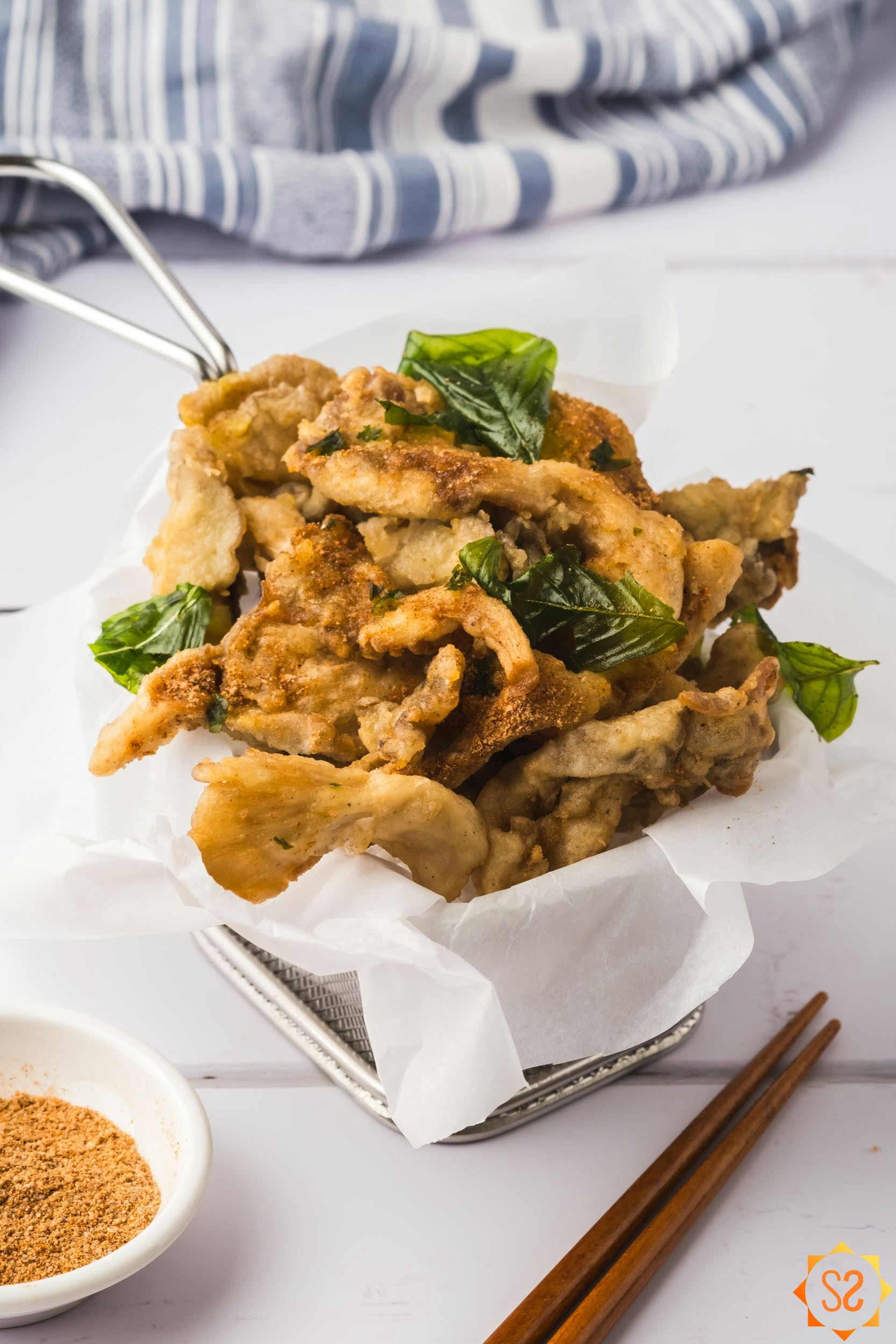 A basket of fried oyster mushrooms with seasoning powder and chopsticks to the sides. 
