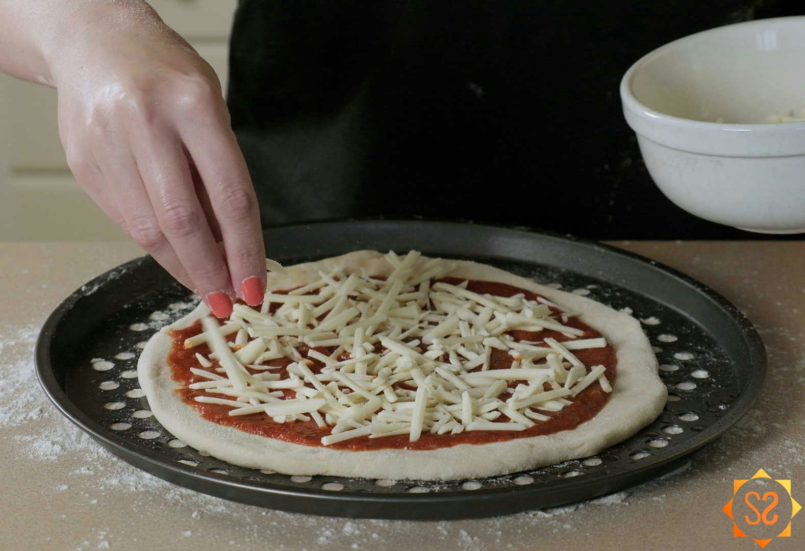 A hand sprinkling vegan cheese on top of an uncooked pizza on a vented pizza pan.