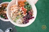 A top-down view of a Buddha bowl with quinoa, vegetables, and hummus, being held in a person's hand.