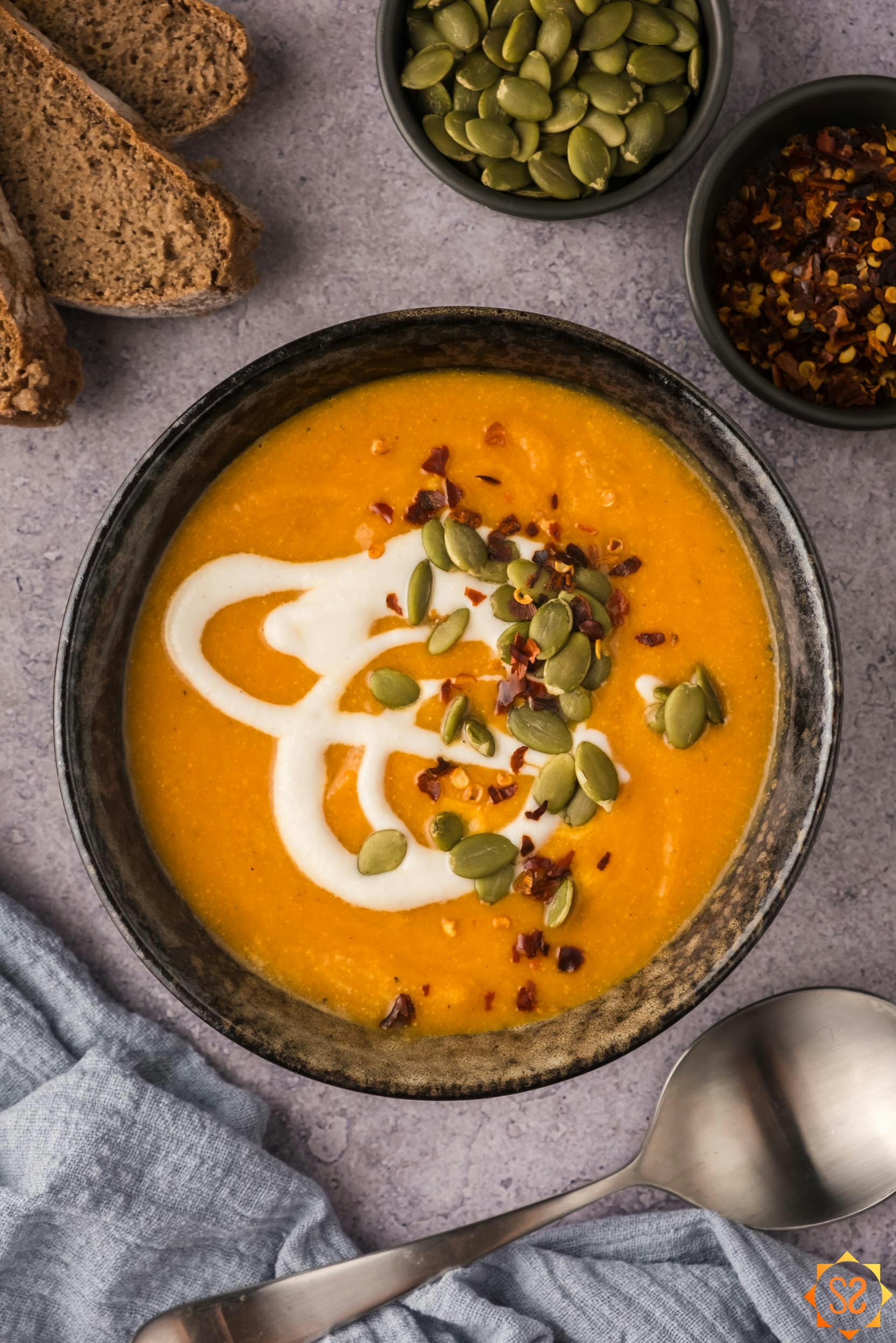 A top-down view of a bowl of carrot pumpkin soup surrounded by bread and toppings.