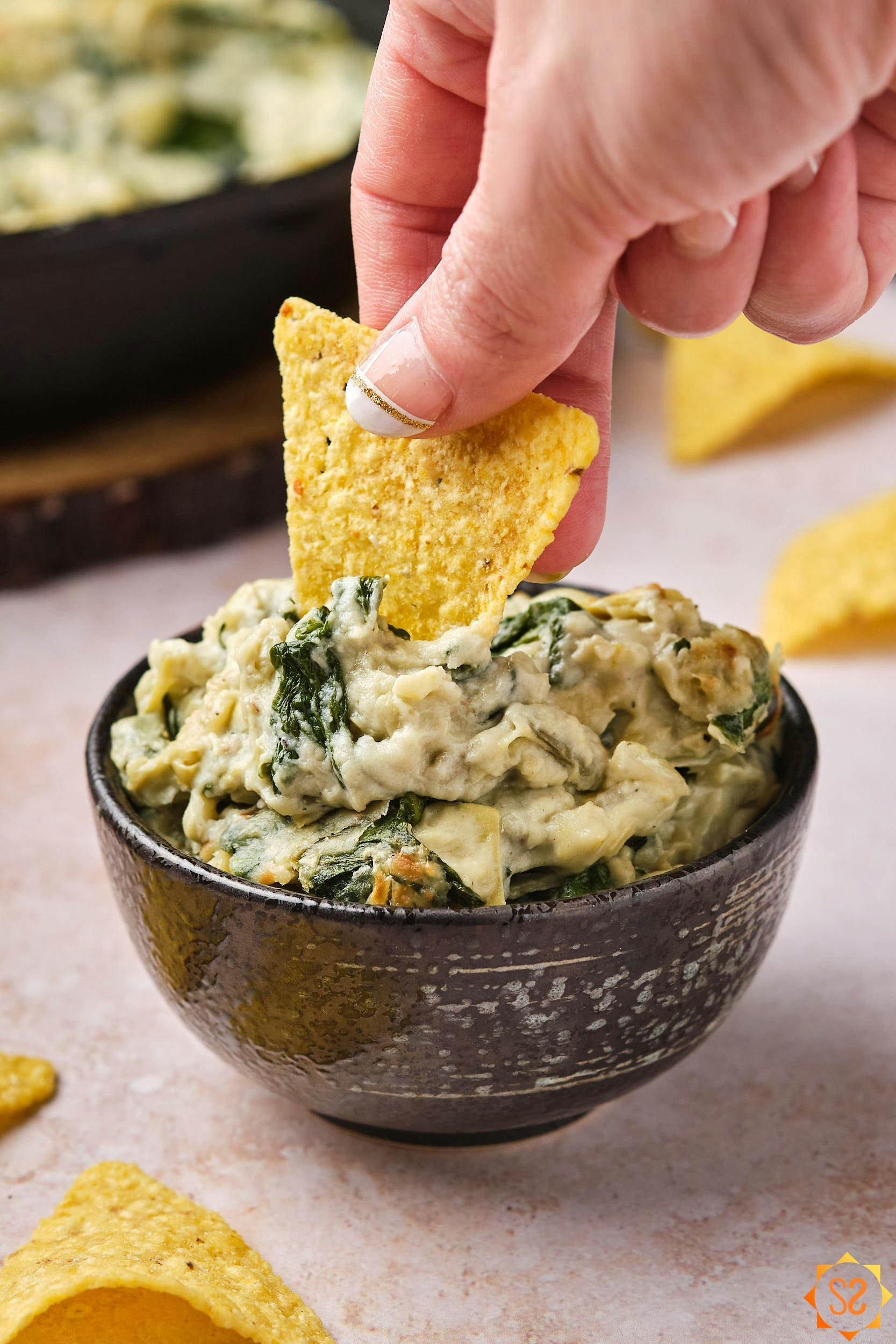 A hand dipping a tortilla chip into a bowl of vegan spinach and artichoke dip. 