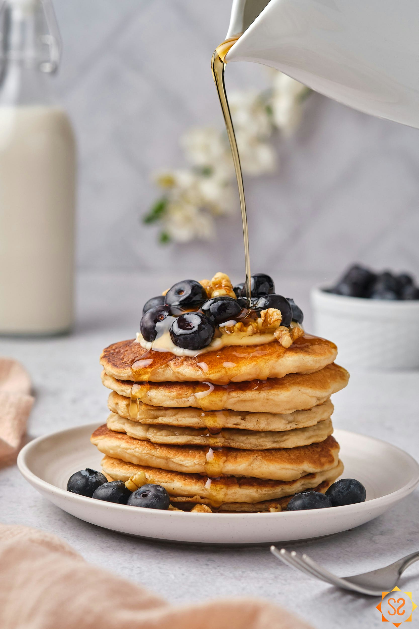 A wide view of a stack of vegan protein pancakes, with maple syrup being poured from a small pitcher above.