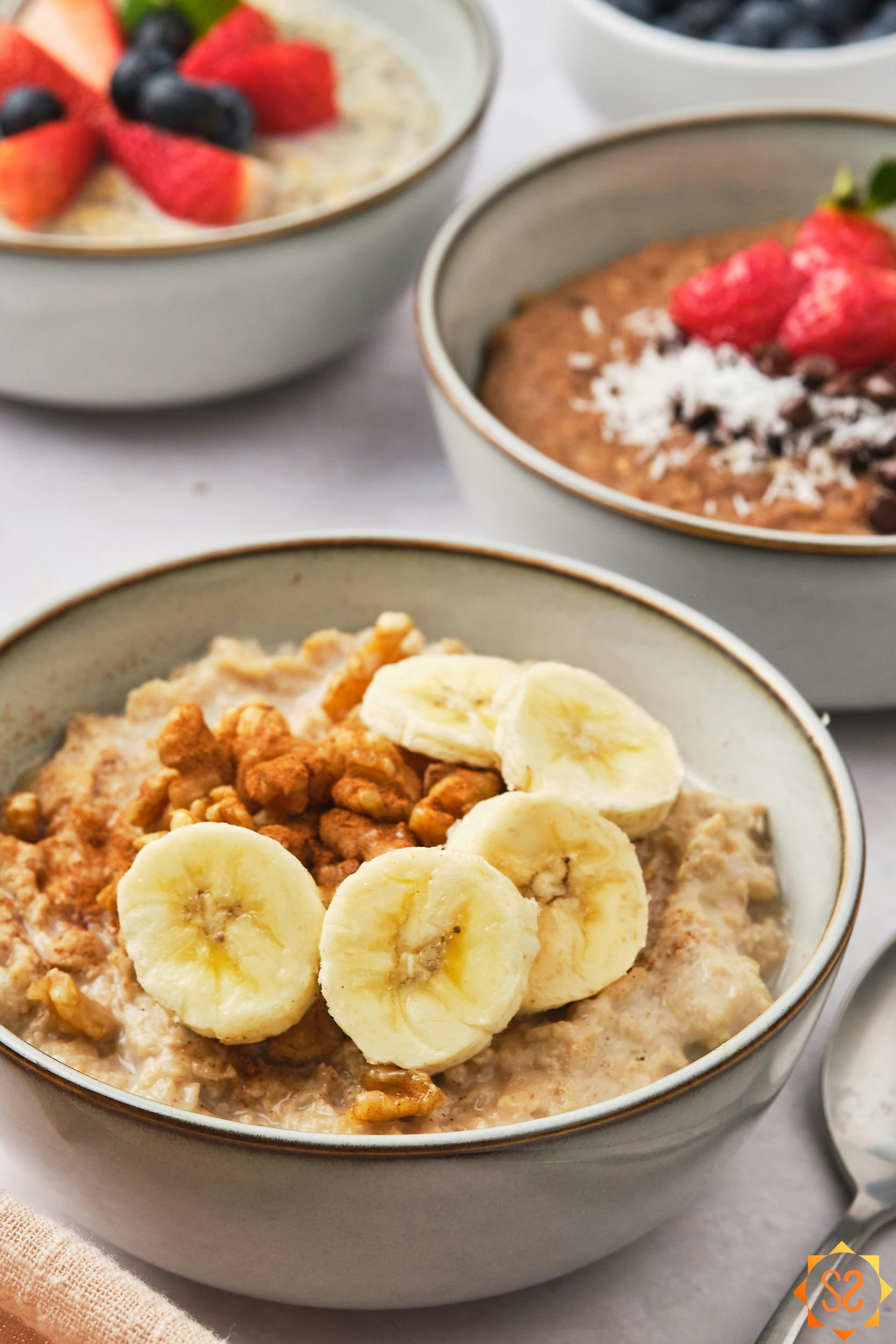 A 45 degree view of three bowls of protein oatmeal, the one in front is topped with banana slices and walnuts.